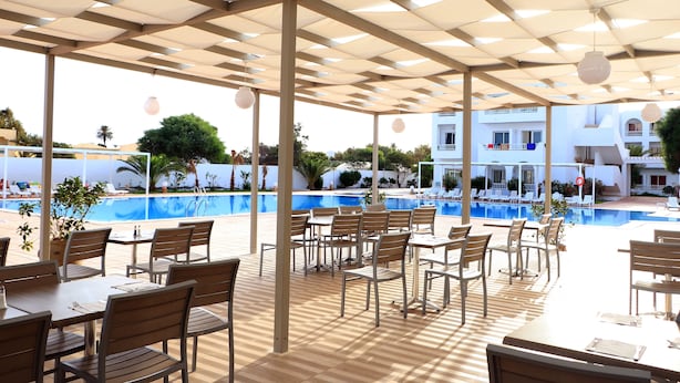 Un espacio de estar al aire libre cubierto, con mesas y sillas, una piscina y un edificio al fondo.