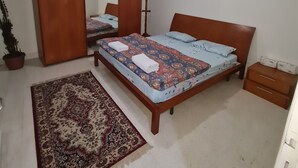 Un dormitorio con una cama, una mesita de noche, un aparador, un espejo, un perchero, una planta en maceta y una alfombra.