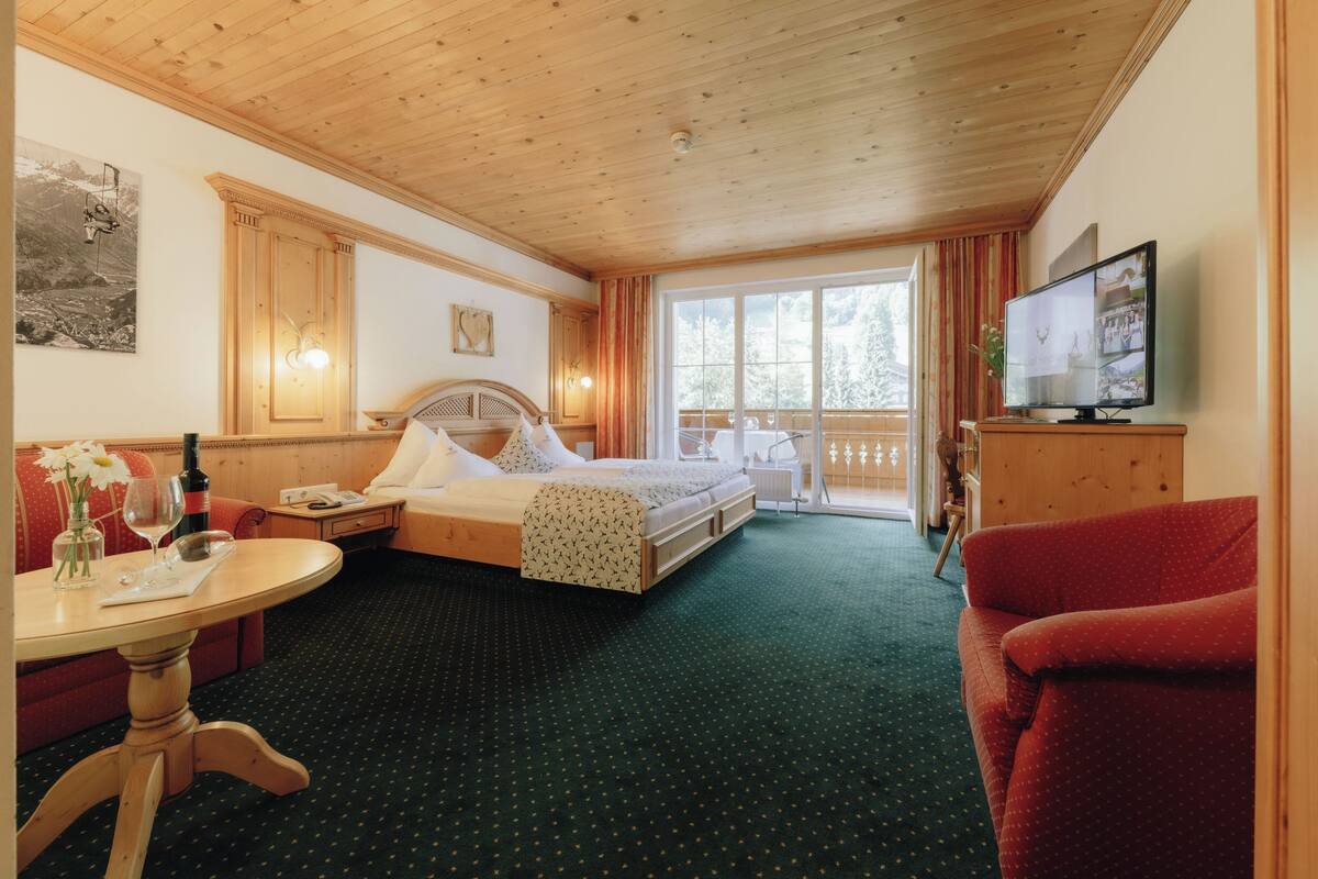 A hotel room with a bed, a television, a dining table, and a balcony with a view of the mountains.