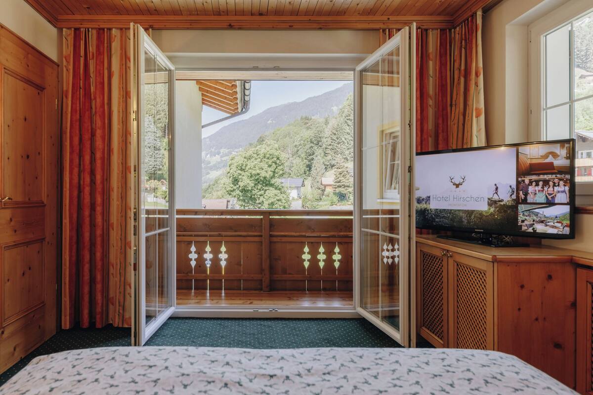 A hotel room with a large sliding glass door leading to a balcony, a bed with a patterned bedspread, and a television displaying a hotel image.