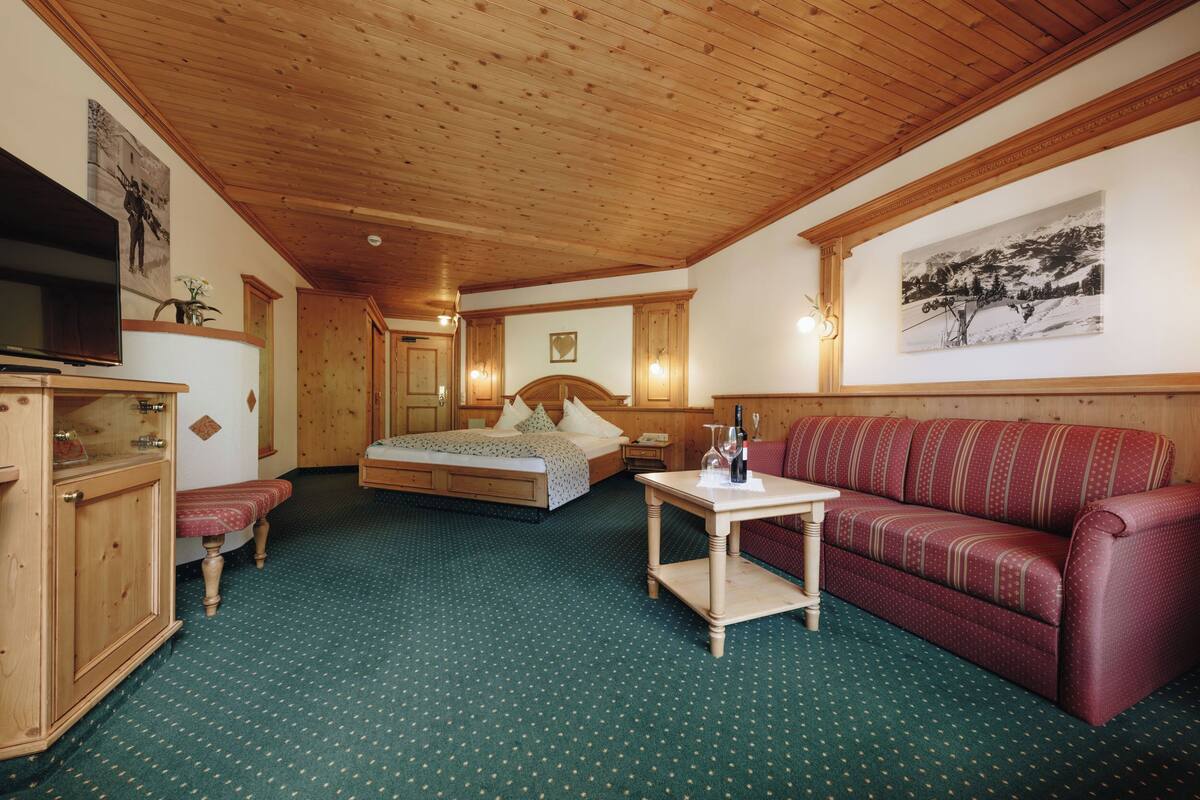 A room with a wooden ceiling, a bed with a headboard, a red sofa, a small table with a bottle, and a television on a cabinet.