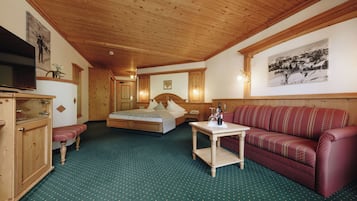 A room with a wooden ceiling, a bed with a headboard, a red sofa, a small table with a bottle, and a television on a cabinet.