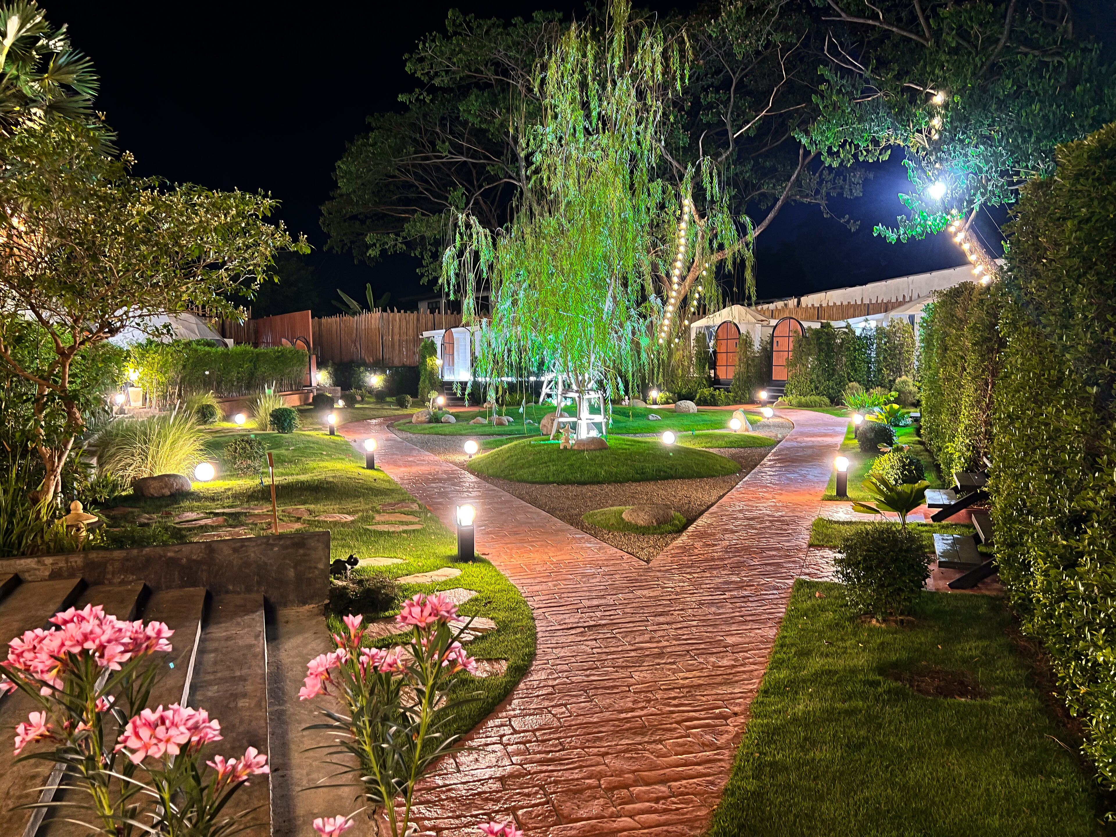 A well-lit garden pathway leading to a building with a wooden fence and a large tree.