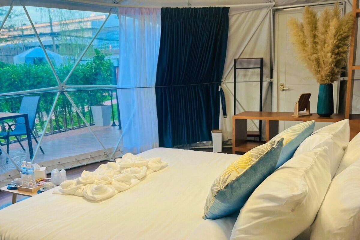 A bedroom with a bed, pillows, a bedside table, a window with curtains, and a view of greenery outside.