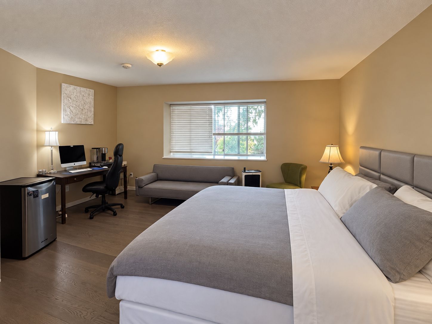 A hotel room with a bed, a desk with a computer, a chair, and a window with blinds.