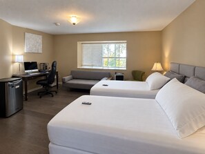Habitación de hotel con dos camas, un escritorio con computadora, una silla, un sofá y una ventana con persianas.