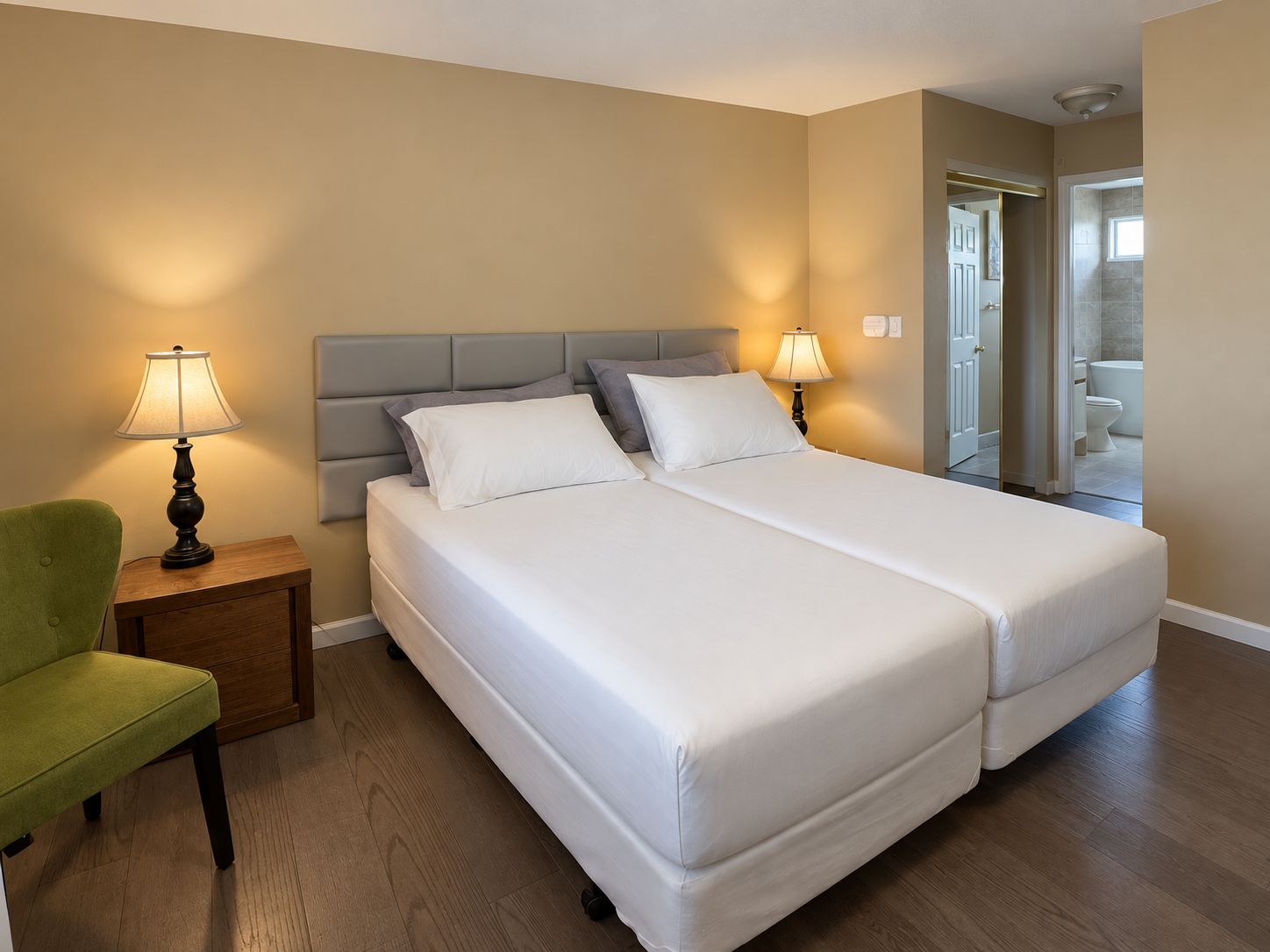 A hotel room with a double bed, two bedside lamps, a green armchair, and a bathroom visible through an open door.