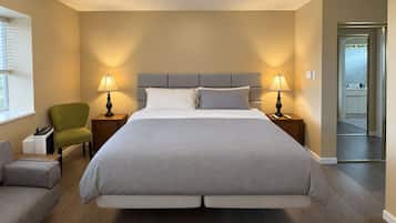 A hotel room with a large bed, two bedside tables with lamps, a green armchair, a grey sofa, and a view of the sea through a glass door.