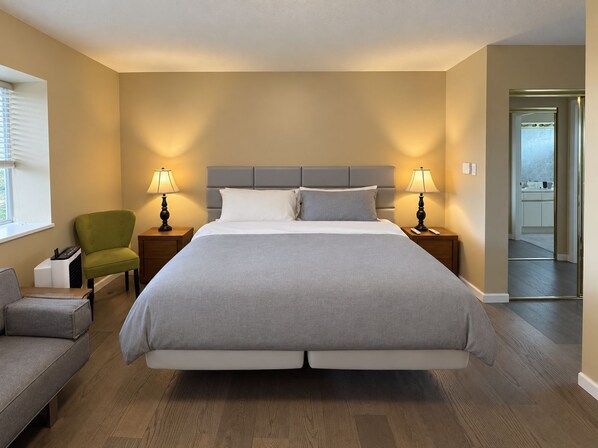 Habitación de hotel con una cama grande, dos mesitas de noche con lámparas, un sillón verde, un sofá gris y vista al mar a través de un ventanal.