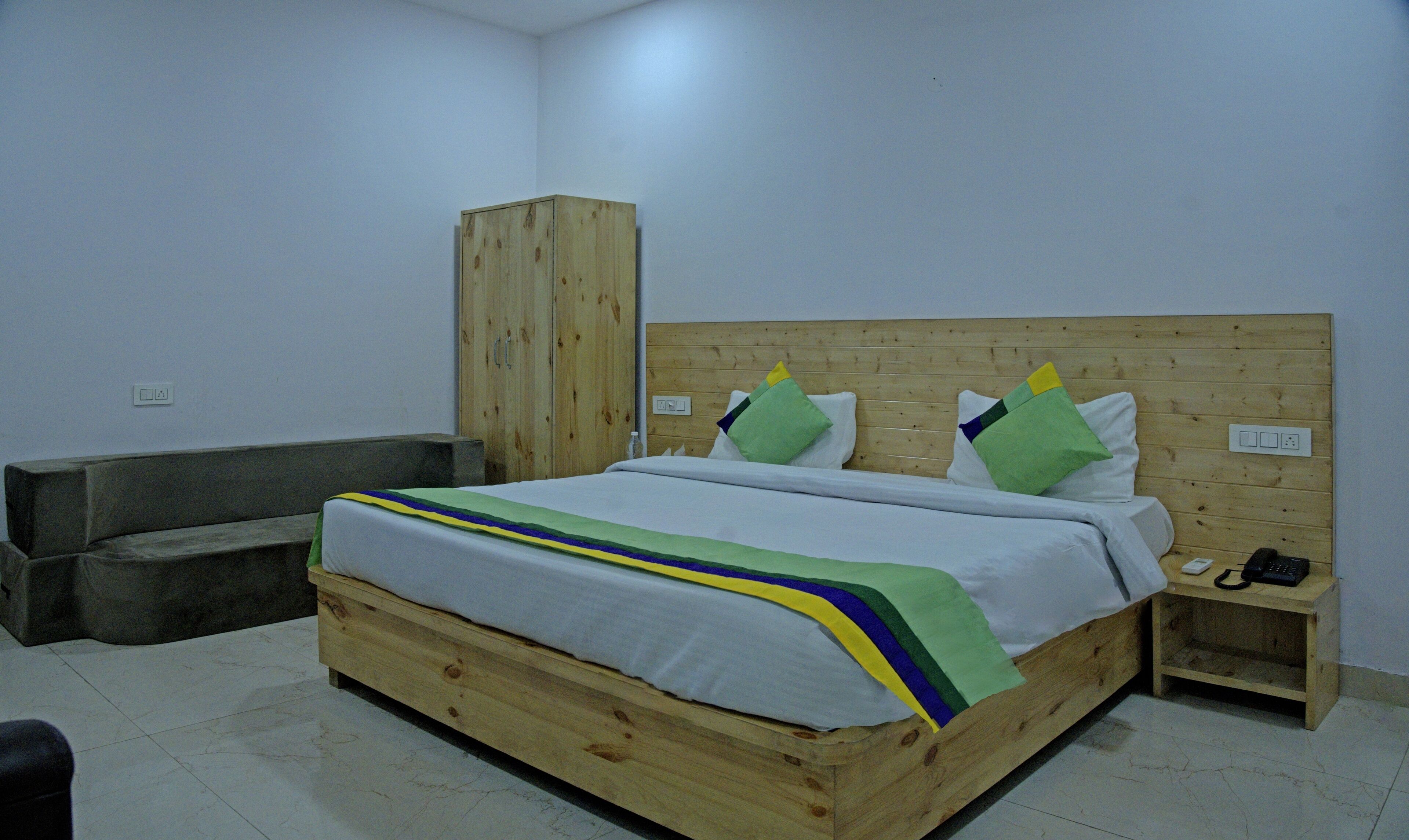 Een houten bed met een witte matras, een blauw-groen gestreept dekbed en meerdere kussens. Daarbij een houten garderobe en een klein nachtkastje met een telefoon en een afstandsbediening.