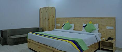 Een houten bed met een witte matras, een blauw-groen gestreept dekbed en meerdere kussens. Daarbij een houten garderobe en een klein nachtkastje met een telefoon en een afstandsbediening.