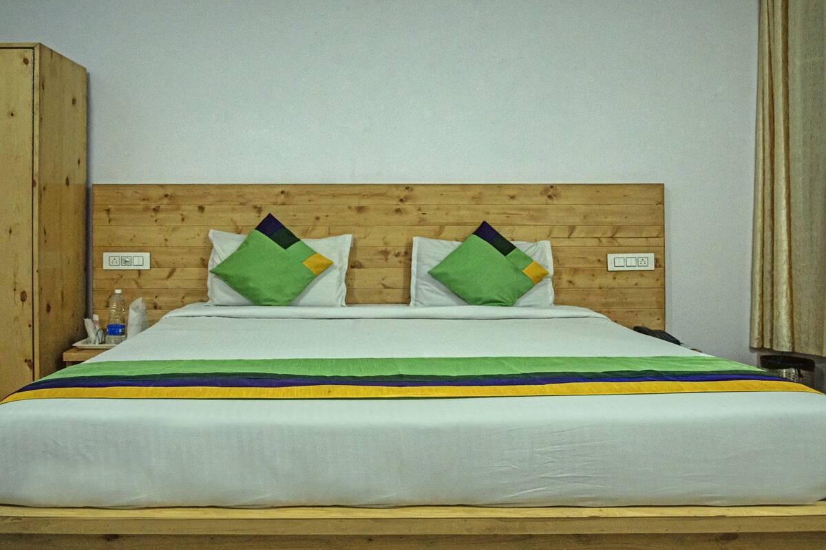 A bed with a wooden headboard, two pillows, a striped blanket, and a wooden nightstand with a bottle and a remote control.
