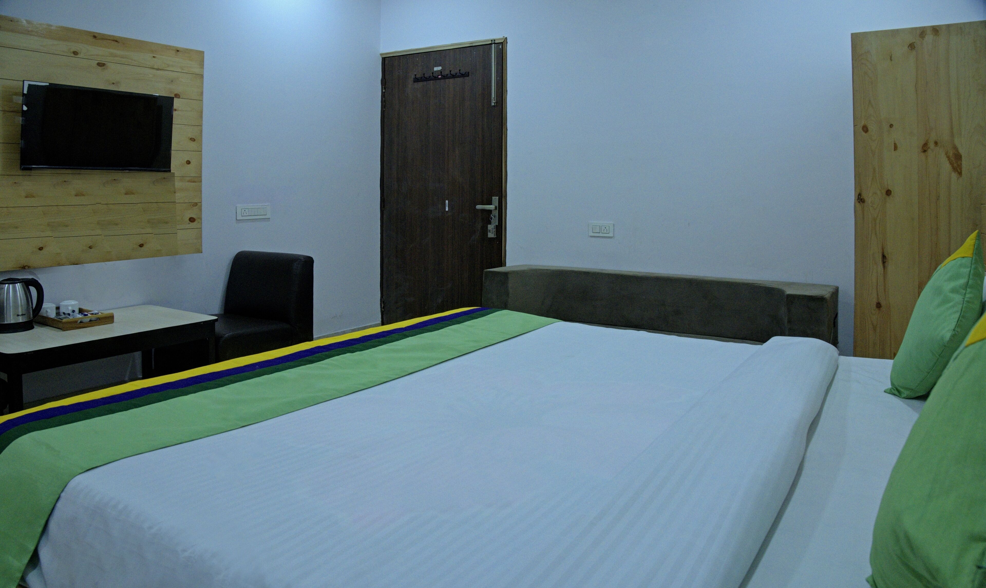 A hotel room with a bed, a desk with a chair, a television mounted on the wall, and a wooden door.