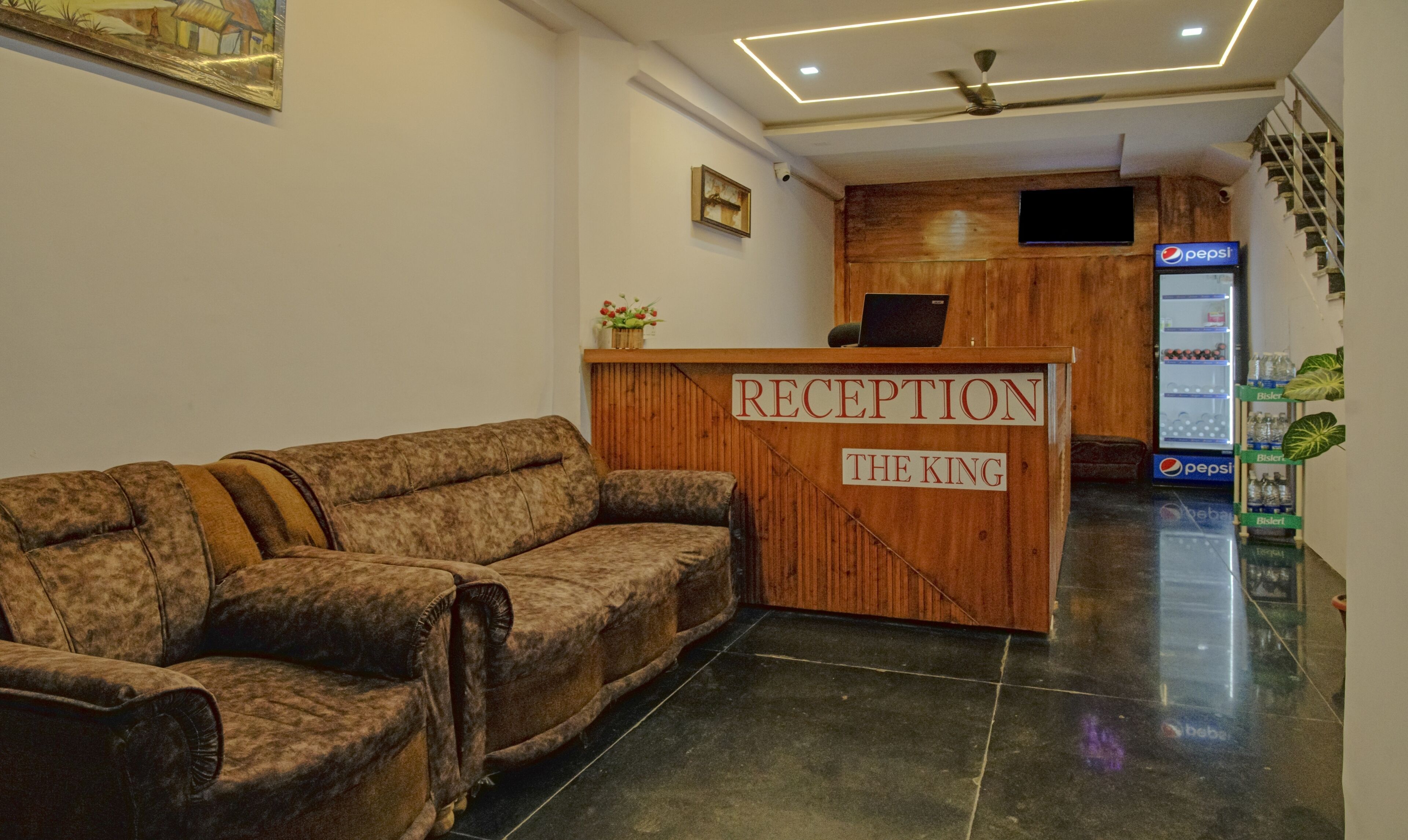 Ein Empfangsbereich mit einem hölzernen Empfangstresen mit der Aufschrift "RECEPTION THE KING", einer Couch, einem Deckenventilator, einem an der Wand montierten Fernseher und einem Kühlschrank für Pepsi.