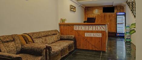 A reception area with a wooden reception desk labeled "RECEPTION THE KING," a sofa, a ceiling fan, a wall-mounted TV, and a Pepsi refrigerator.