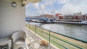 A balcony with wicker furniture, a view of a river, a bridge, and buildings in the background. - 2 Bedroom River Front Condo (Manistee)