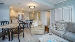 A modern living room with a sofa, armchair, and coffee table. There is a dining area with a table and chairs. The kitchen is visible in the background with cabinets and appliances. - 2 Bedroom River Front Condo (Manistee)