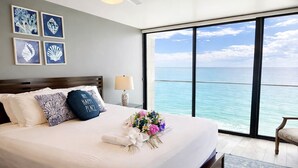 A hotel room with a large bed, a nightstand, a lamp, and a view of the ocean. - Ocean View Luxury Condo: Beachfront, and Pool (San Diego)