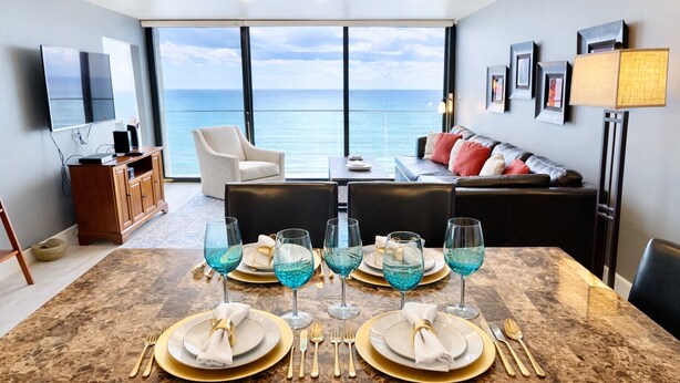 A dining area with a table set for four, a sofa, and a view of the ocean. - Ocean View Luxury Condo: Beachfront, and Pool (San Diego)