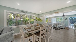 A dining area with a large table set for four, surrounded by white chairs. There is a large window with a view of a wooden fence and greenery. A ceiling fan is visible in the dining area. - 7 minute walk to Siesta Key beach.  Large heated pool/spa. Very private.1/2 acre (Siesta Key)