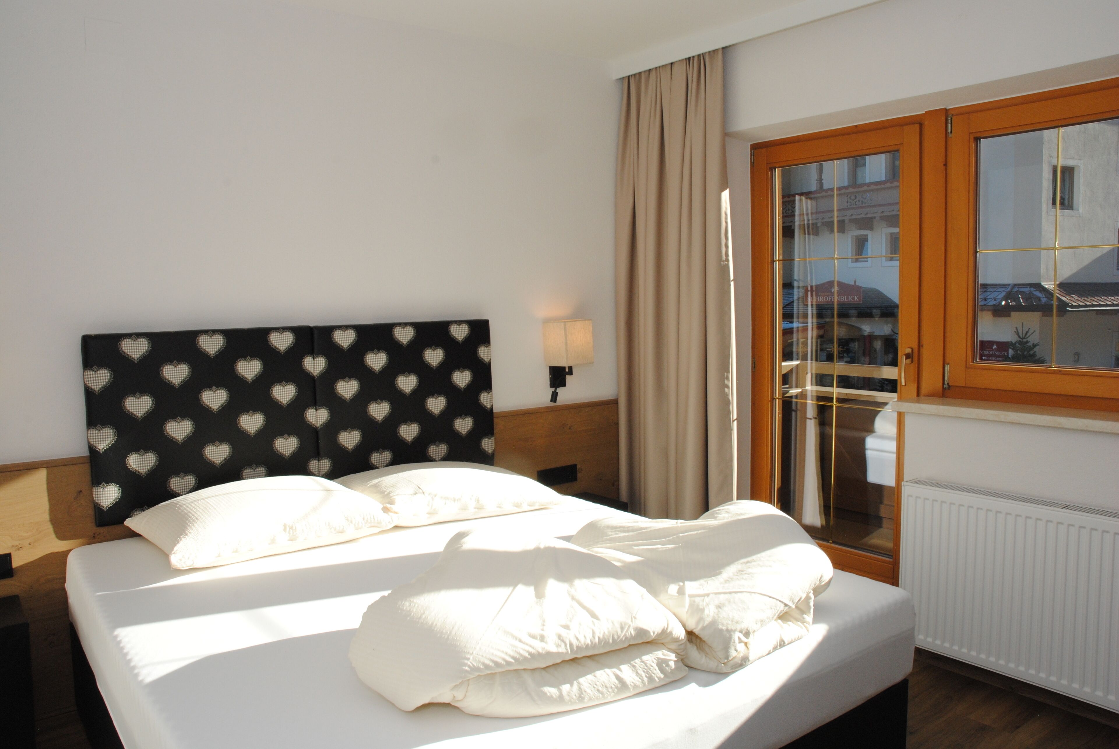 A hotel room with a bed, a headboard with a heart pattern, a nightstand with a lamp, a window with a view of buildings, and a radiator.
