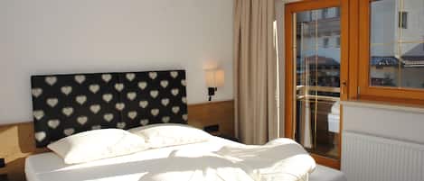 Una habitación de hotel con cama, cabecera con estampado de corazón, mesita de noche con lámpara, ventana con vistas a edificios y un radiador.