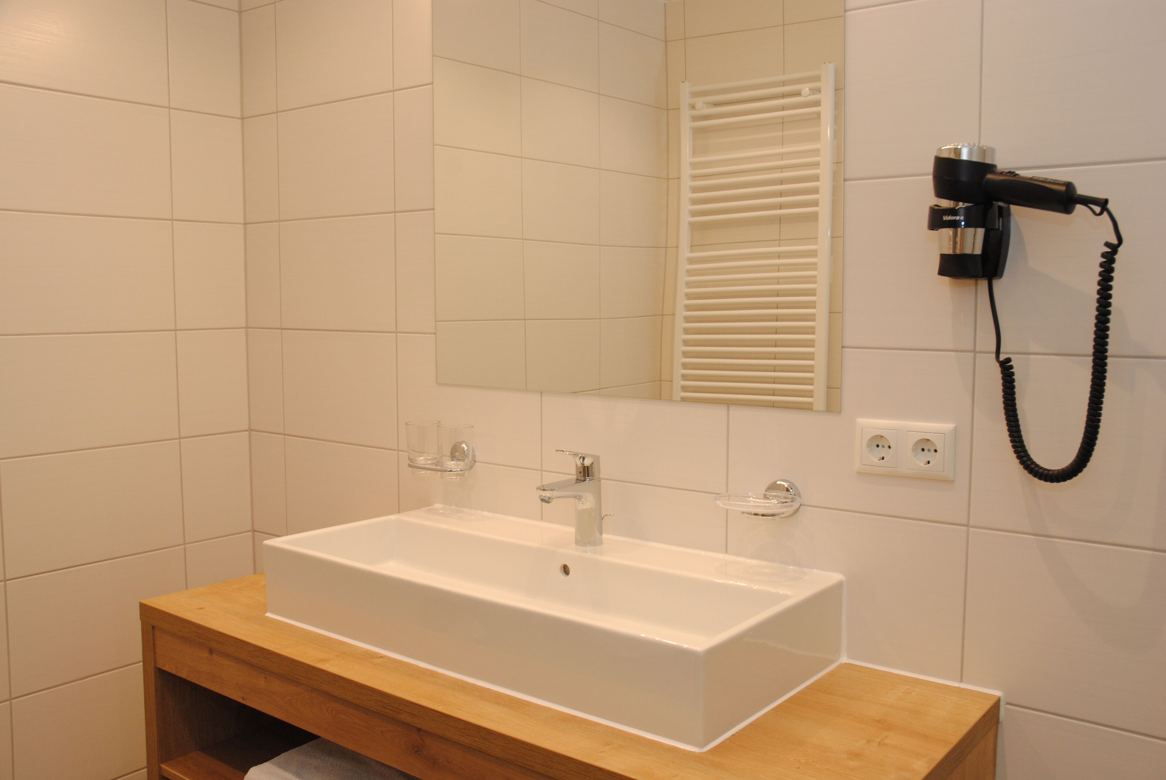 Een badkamer met een witte wastafel, een houten wastafelmeubel en een haardroger bevestigd aan de muur.