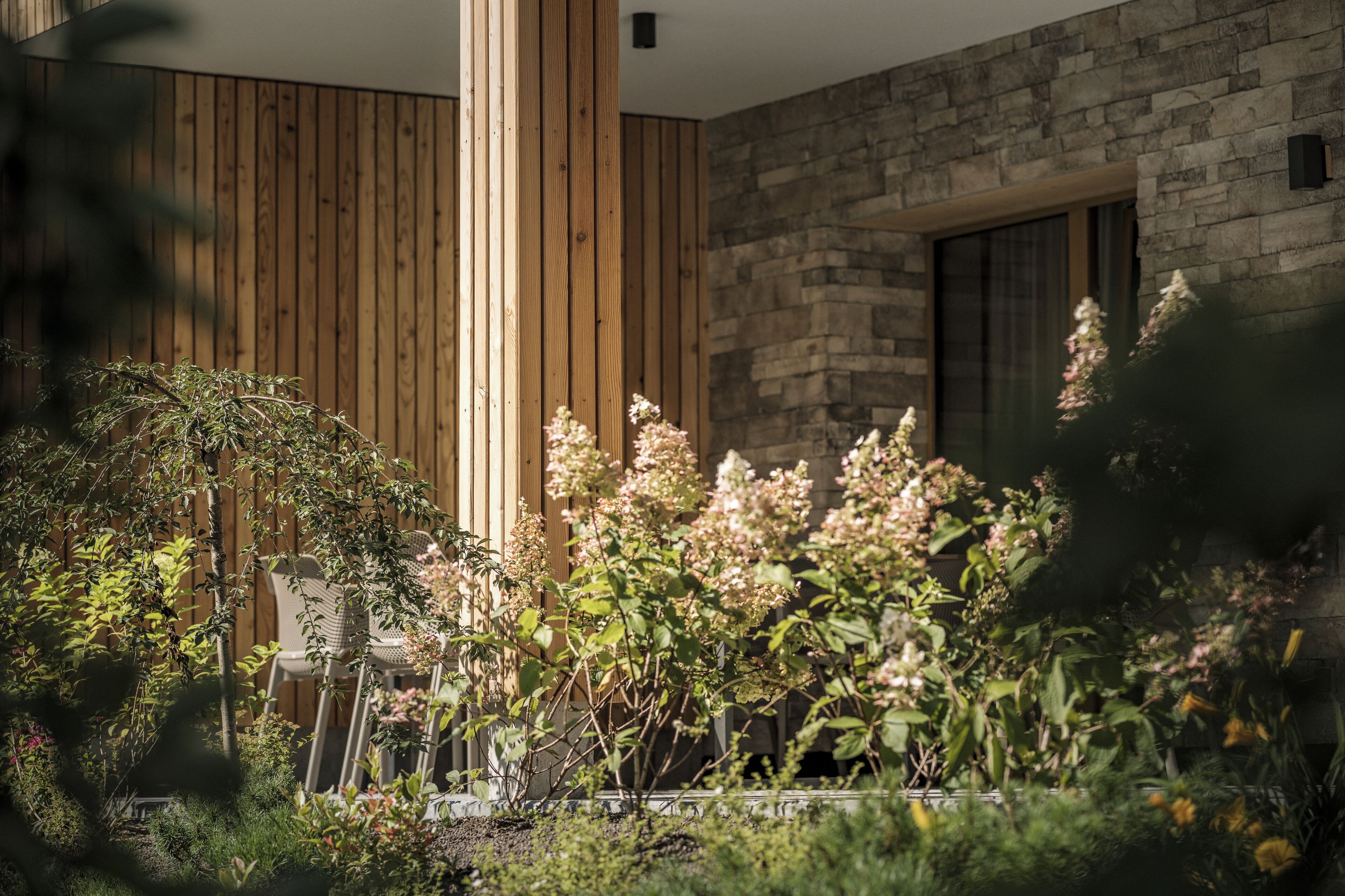 A modern building with wooden paneling and stone walls, surrounded by lush greenery and flowering plants.