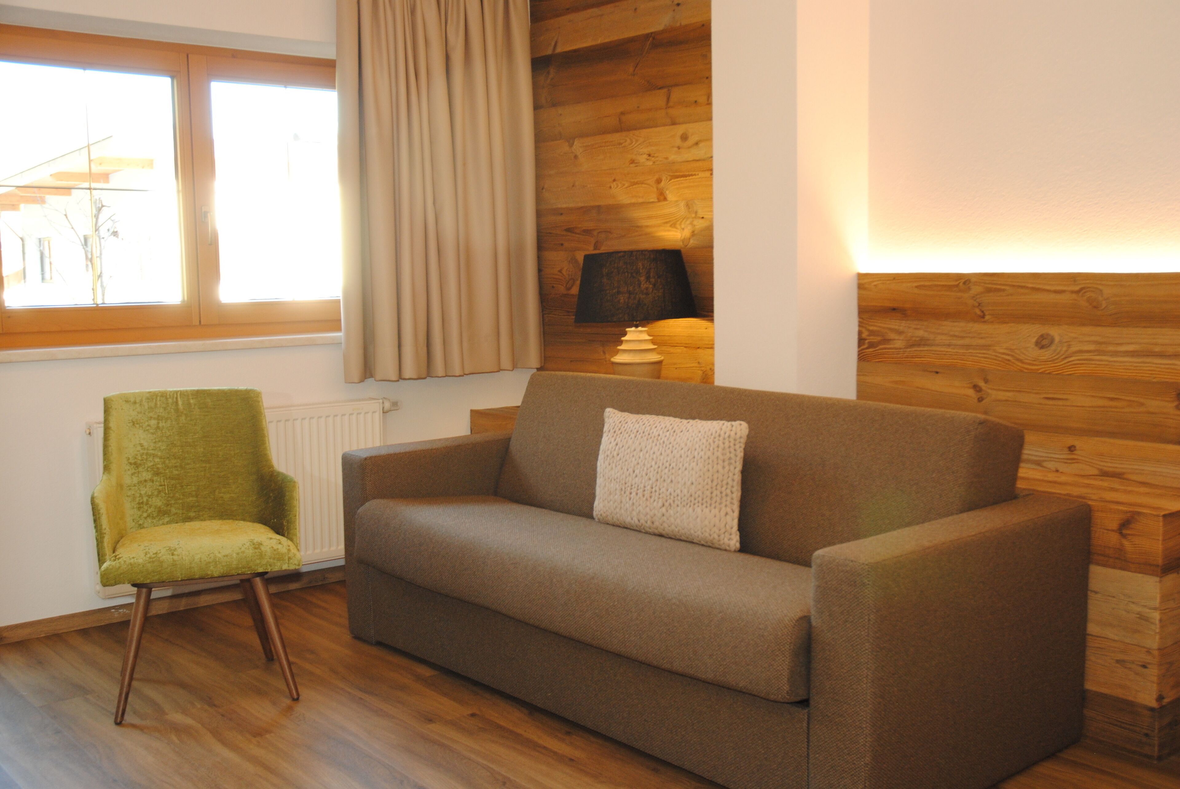 Ein Zimmer mit Holzboden, einem grünen Sessel, einem braunen Sofa mit Kissen, einer Lampe und einem Fenster mit Vorhängen.