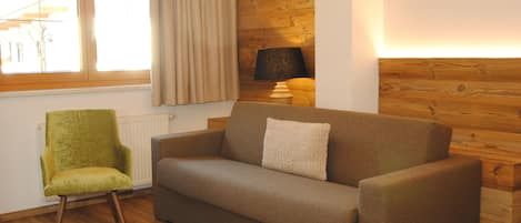 Une pièce avec un plancher en bois, une chaise verte, un canapé marron avec un coussin, une lampe et une fenêtre avec des rideaux.