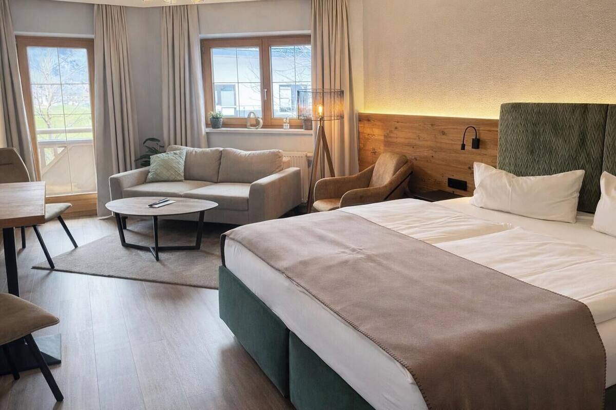 Une chambre d’hôtel avec un grand lit, deux fauteuils, une petite table ronde et une vue sur l’extérieur grâce à de grandes fenêtres.