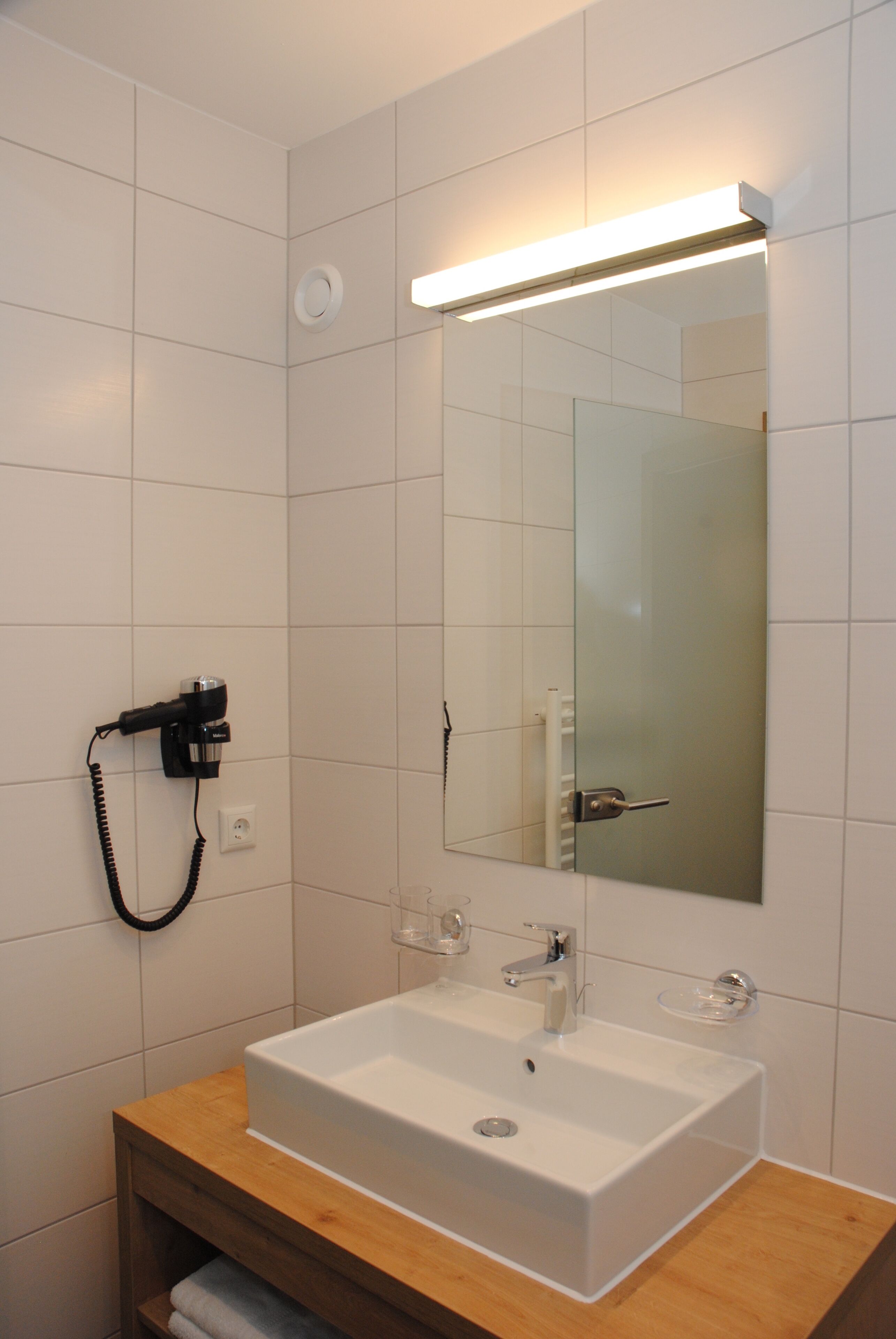 Een moderne badkamer met een witte wastafel, spiegel en een haardroger aan de muur.