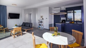 Una sala moderna con un sofá, sillas, una mesa de comedor con cojines amarillos, un televisor de pantalla plana en la pared y una zona de cocina con armarios azules y una ventana.