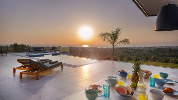 Una terraza soleada con sillones de descanso, una piscina y una mesa dispuesta con alimentos para el desayuno.