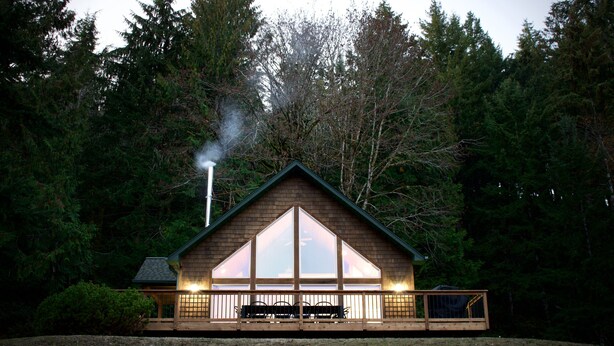 A cabin with a large window, a deck, and a chimney emitting smoke.