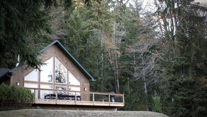 A house with a large deck and a sloped roof, surrounded by dense forest.