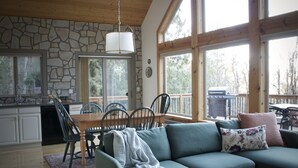 A cozy living room with a green sofa, a dining table with chairs, a kitchen area with a dishwasher, and large windows offering a view of the outdoors.