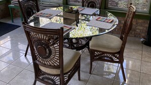 A round glass table with four wicker chairs, surrounded by large windows offering a view of trees and a garden.