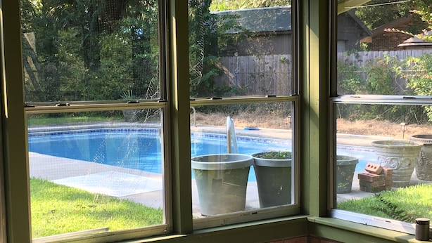 A room with a view of a swimming pool through large windows, a brick wall, and a small stool.
