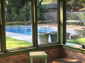 A room with a view of a swimming pool through large windows, a brick wall, and a small stool.