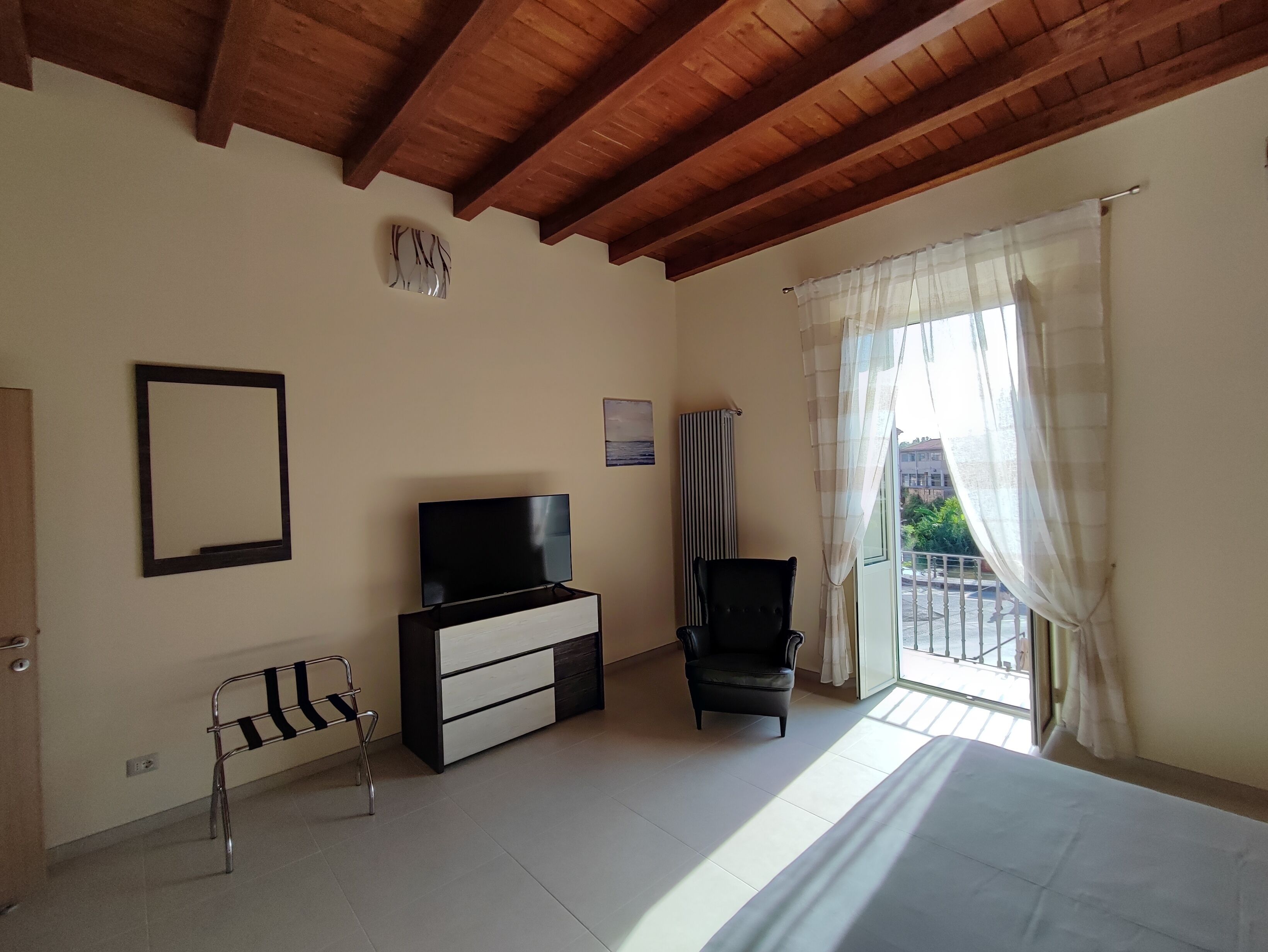 Quarto com cama, televisão, cadeira, radiador e janela com cortinas.