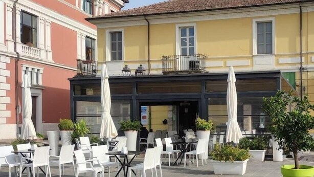 An outdoor cafe with white chairs and tables, potted plants, and a yellow building with a balcony. - In Centro - Rooms and Apartments  (Isola del Liri)