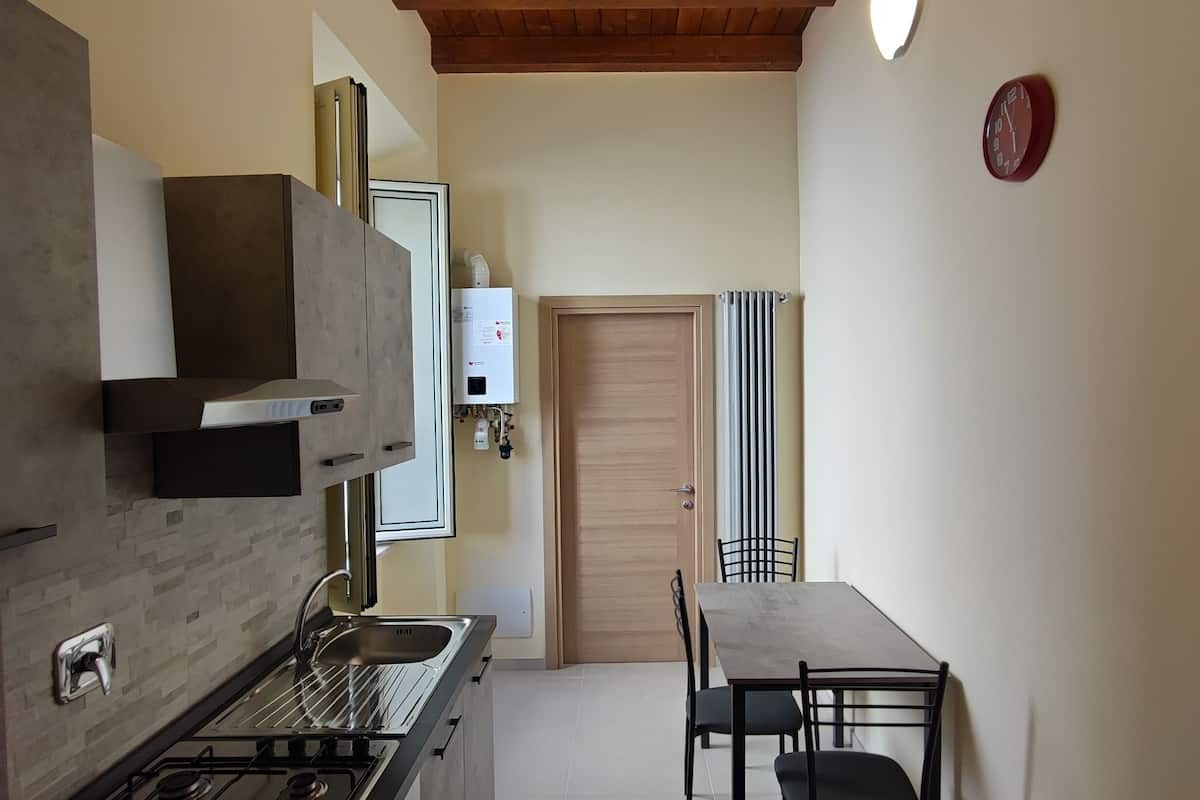 A compact kitchen with a gas stove, oven, and a microwave. There is a dining table with chairs and a red rug. The ceiling is wooden with exposed beams, and there are two wall-mounted lights.