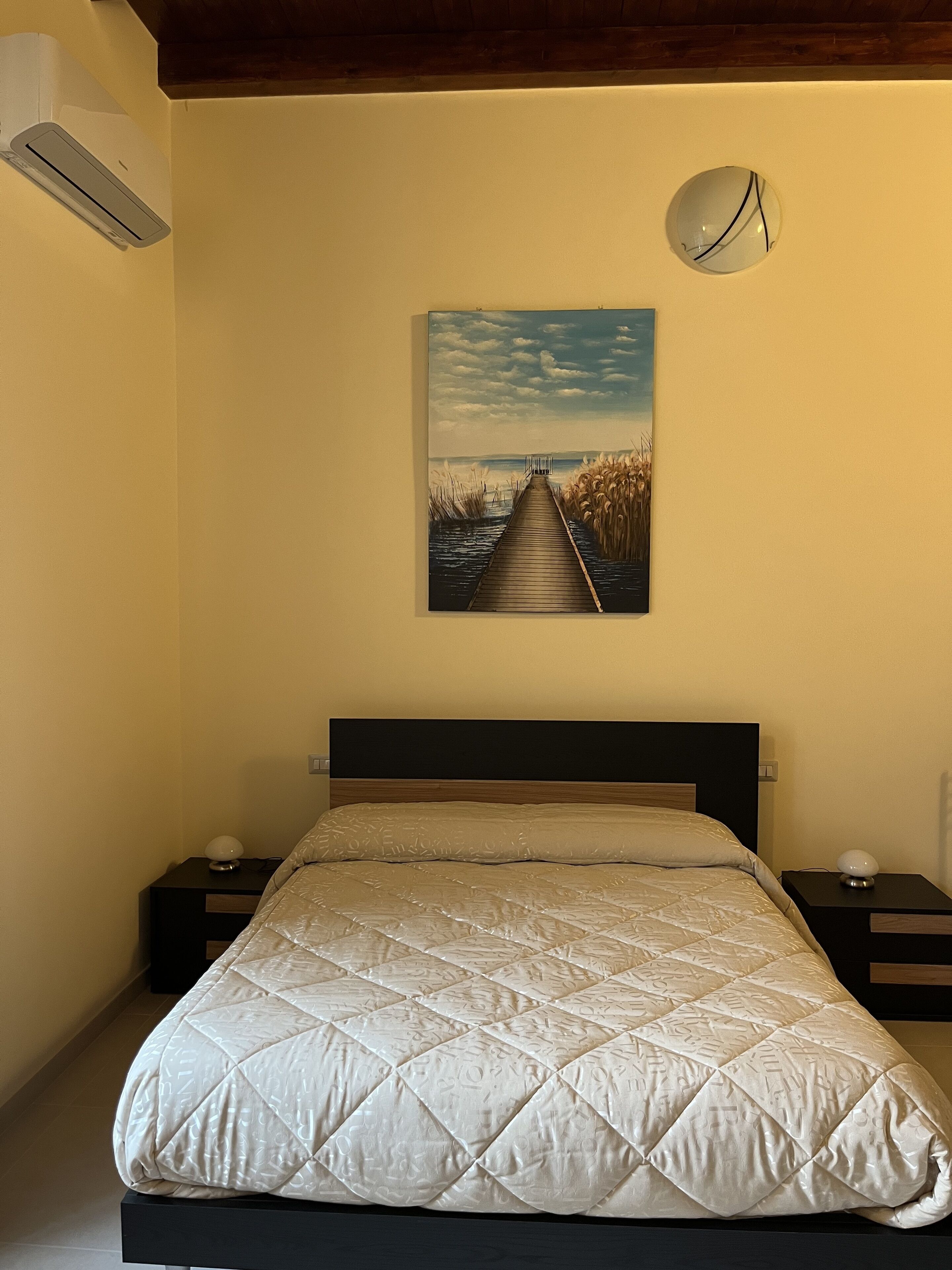 Ein Schlafzimmer mit einem Bett, einem Nachttisch, einer Wand-Klimaanlage und einem Gemälde eines Piers.