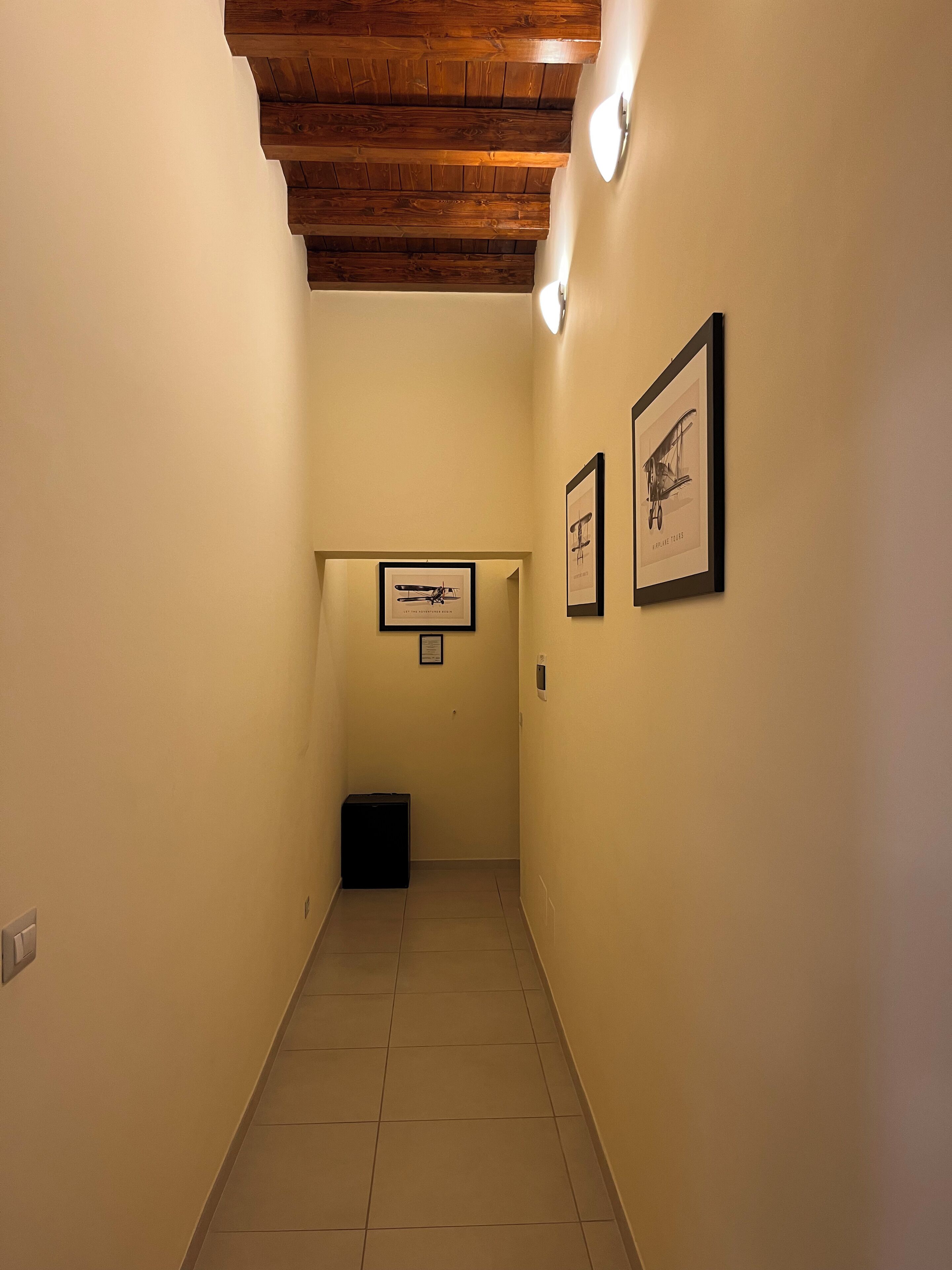 A narrow hallway with wooden ceiling beams, tiled floor, and framed pictures on the walls.