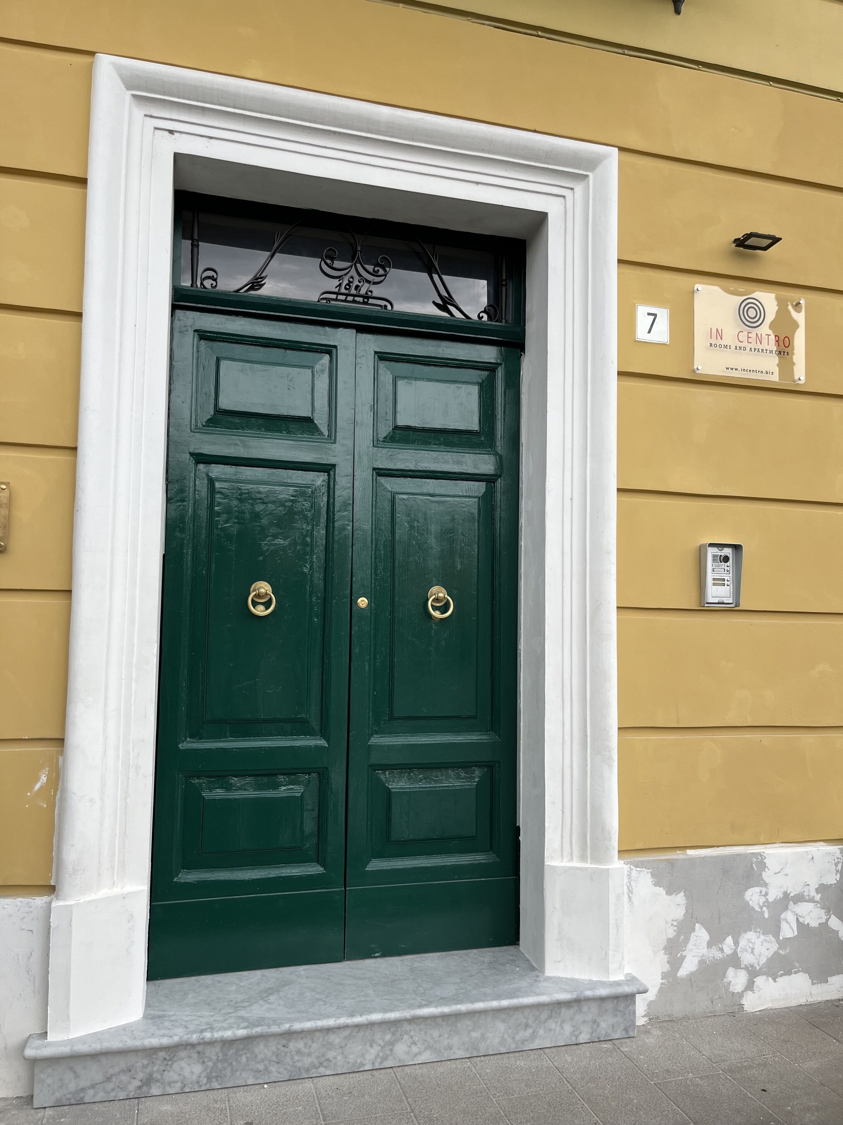 Puerta doble verde con manijas de latón, enmarcada en blanco, sobre una pared amarilla con un letrero que indica "7" y "In Centro Rooms and Apartments".