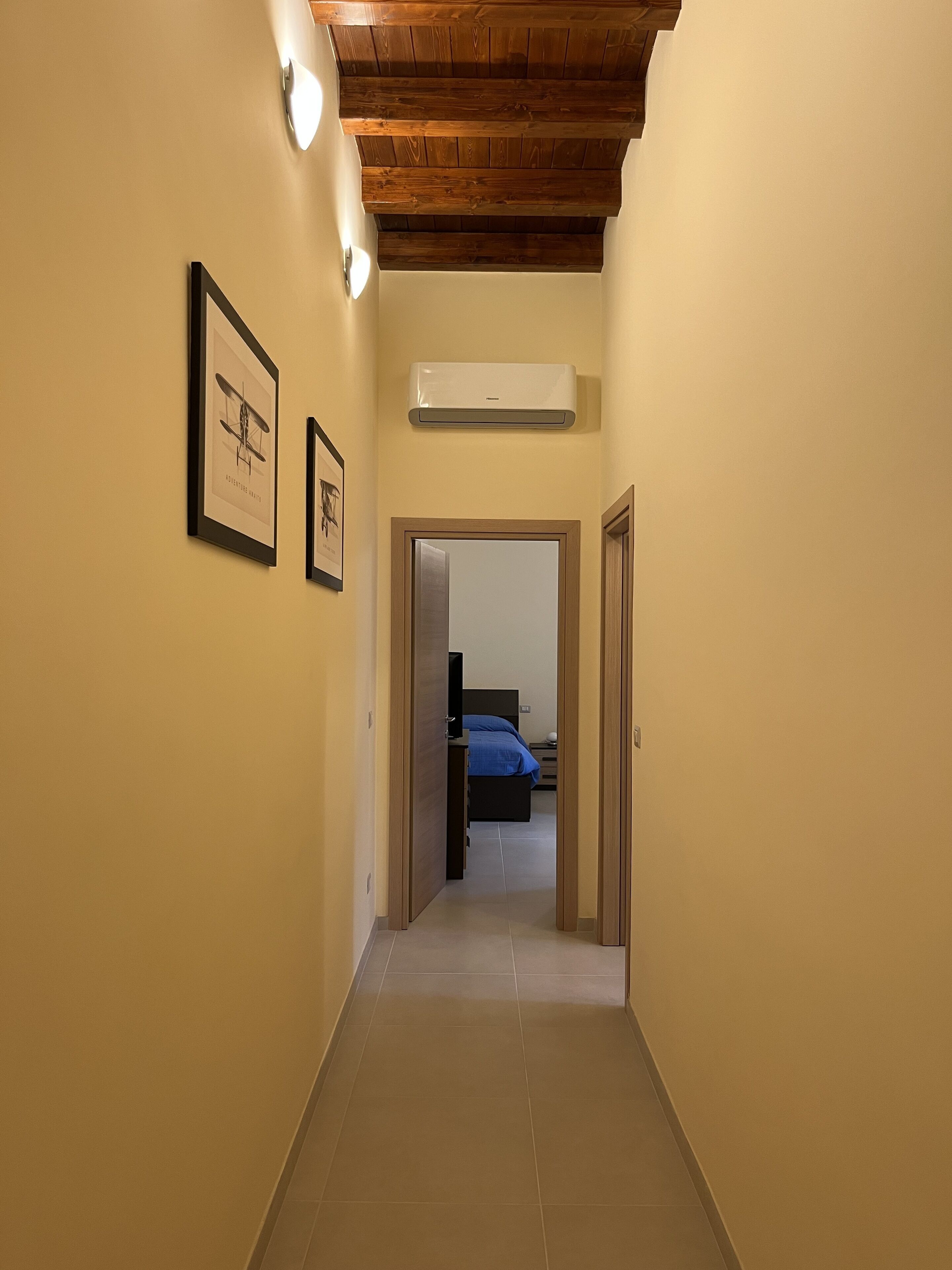A hallway with a wooden ceiling, framed pictures on the walls, and a bed in the background.