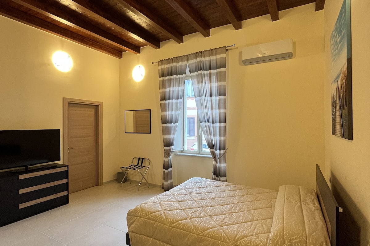A bedroom with a bed, a TV, a dresser, a window with curtains, and an air conditioning unit.