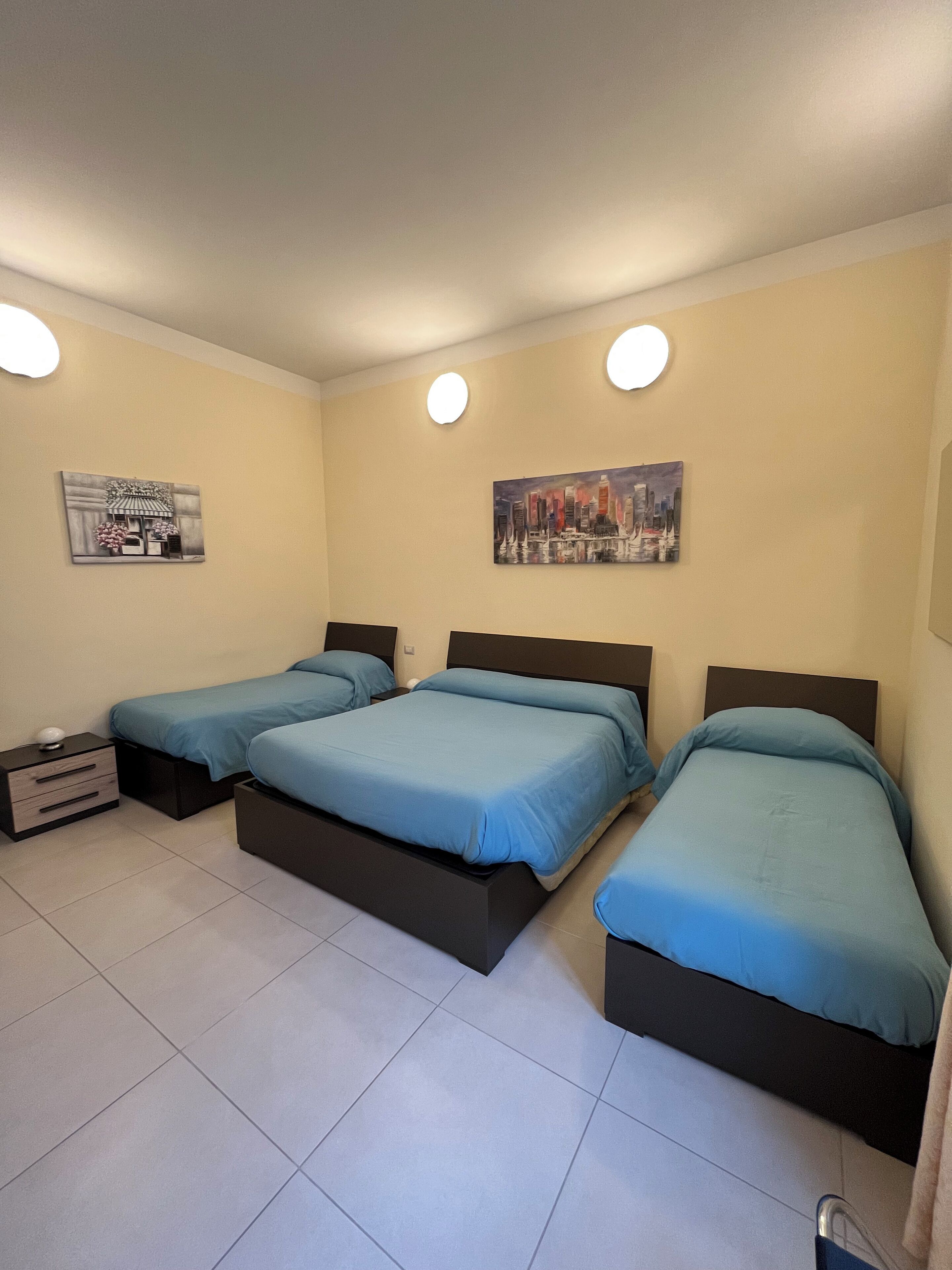 A bedroom with three beds, each with a blue mattress and black headboard. There are two wall-mounted light fixtures and two paintings.