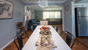A dining area with a long table set for four, a chandelier, and a modern kitchen with a refrigerator. - Beautiful Spacious Home- Near the Expo, TU, gathering Place, Fair park (Tulsa)
